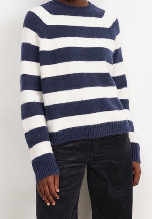 Personne portant un pull à rayures horizontales bleu marine et blanc avec des poignets côtelés et un pantalon en velours côtelé foncé.