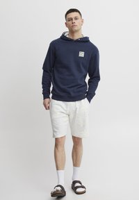 Navy blue hooded sweatshirt with a front patch logo, paired with light grey shorts. The outfit is complemented by brown sandals and white socks.
