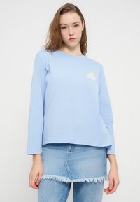 Ανοιχτό μπλε μακρυμάνικο πουκάμισο από βαμβάκι, με μικρό κίτρινο και λευκό γραφικό σχέδιο στο στήθος και χαλαρή γραμμή.