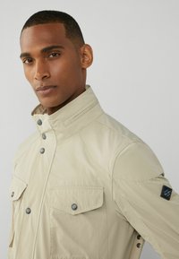 Beige lättviktsjacka med hög krage, tryckknappar, två bröstfickor och en texturerad ärmplatta. Slät tyg med modern passform.