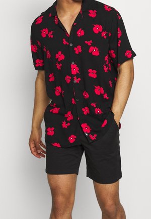 Mand iført sort kortærmet skjorte med knapper og lyst rødt blomsterprint samt sorte shorts, stående med den ene hånd i lommen.