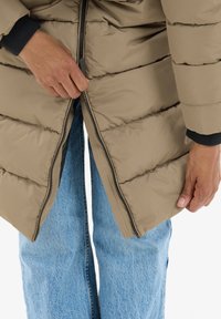Lys brun puffjakke med vandrette quiltede sektioner. Lynlås halvmåne åben, præsenteret mod blå denim jeans. Tekstureret stof, afslappet design.