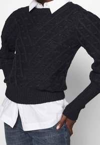 Svart stickad tröja med ett texturerat kabelmönster, ribbade manschetter och nederkant. Lagd över en vit skjorta med krage.