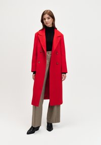 Czerwony płaszcz dwurzędowy z dużymi klapami i złotymi guzikami, zestawiony z czarnym swetrem i szerokimi spodniami w kratę. Czarne botki na obcasie.