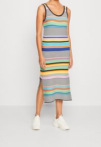 Robe sans manches rayée avec des rayures horizontales multicolores de différentes largeurs, dotée d'une bordure noire et d'une fente latérale, portée avec des baskets blanches.