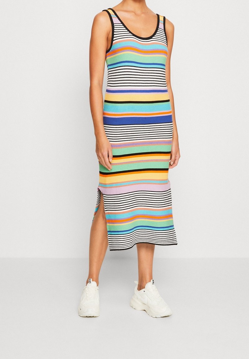 Robe sans manches rayée avec des rayures horizontales multicolores de différentes largeurs, dotée d'une bordure noire et d'une fente latérale, portée avec des baskets blanches.