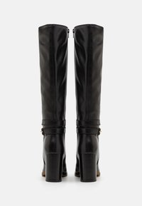 Bottes noires en cuir jusqu'au genou avec une finition lisse, dotées d'un talon carré et d'une sangle décorative avec quincaillerie dorée. Fermeture éclair à l'arrière.