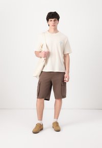 Camiseta de manga corta color beige claro con gráfico azul, combinada con pantalones cortos de cargo marrones con bolsillos laterales. Zapatillas color topo y bolso tote en color blanco roto.