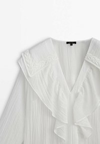 Blouse blanche en tissu texturé, col en V, devant à volants et détail de col carré brodé.