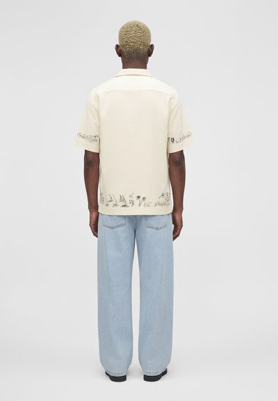 Άτομο με κοντά ξανθά μαλλιά που φοράει μπεζ κοντομάνικο πουκάμισο με μαύρη γραμμική τέχνη και ανοιχτό μπλε φαρδιά τζιν, στέκεται γυρισμένο προς τα πίσω.