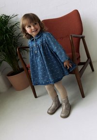 Rifľové šaty s golierom, modré s bielymi vzormi, drevenými gombíkmi a nafúknutými rukávmi. Oblečenie dopĺňajú telové pančuchy a sivé členkové čižmy.