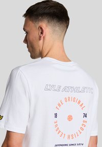 Camiseta de algodón blanca con el texto "LYLE ATHLETIC" y un gráfico circular en naranja y azul marino, ubicado en la parte posterior, con un parche de logo en la manga.