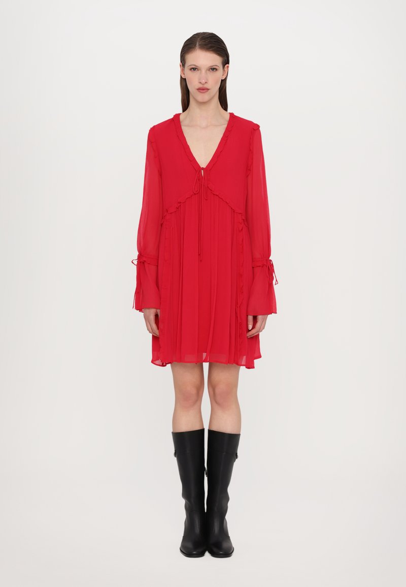 Femme debout portant une robe rouge à manches longues avec un décolleté en V profond et des bottes noires midi contre un fond clair.