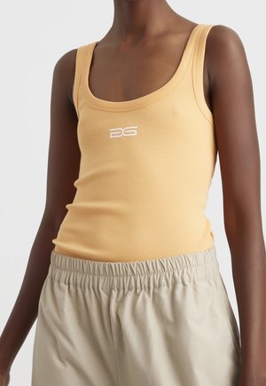 Zbliżenie osoby noszącej beżowe spodnie z elastycznym paskiem oraz żółty prążkowany, bezrękawnik z białym logo na klatce piersiowej.