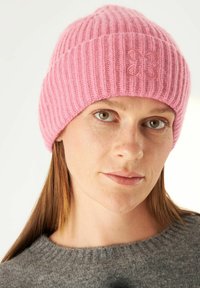 Roze gebreide beanie met verticale ribbels, voorzien van een subtiel vlinderlogo, gedragen met een grijze trui. De textuur lijkt zacht en comfortabel.