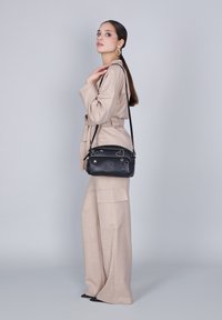 Vrouw in een beige pak en zwarte puntenlaarzen, die een zwarte schoudertas met decoratieve studs draagt, staande tegen een effen grijze achtergrond.
