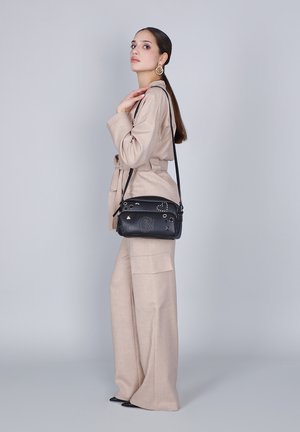 Vrouw in een beige pak en zwarte puntenlaarzen, die een zwarte schoudertas met decoratieve studs draagt, staande tegen een effen grijze achtergrond.