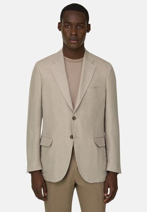 Homme portant un blazer beige à deux boutons sur une chemise beige clair et un pantalon fauve, debout devant un fond uni clair.