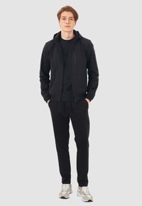 Felpa nera con zip e colletto alto, tasca con zip e pantaloni della tuta neri abbinati. Il modello indossa scarpe da ginnastica chiare. Texture liscia.