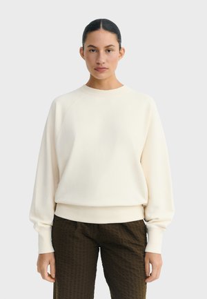 Žena se staženými tmavými vlasy, oblečená v jednobarevné krémově bílé mikině a texturovaných tmavě hnědých kalhotách, stojící před světlým pozadím.