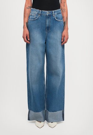 Person, der hochgeschnittene weitbeinige blaue Jeans mit aufgerollten Säumen und weißen spitzen Schuhen trägt, mit tätowierten Armen, entspannt an den Seiten.