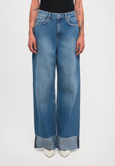 Person wearing high-waist wide-leg blue jeans with rolled cuffs and white pointed-toe shoes, arms tattooed and relaxed by sides.