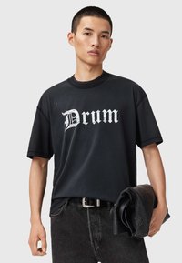 Czarna, oversize koszulka z krótkim rękawem z białą, gotycką grafiką "Drum", z okrągłym dekoltem, w połączeniu z ciemnymi dżinsami i trzymanym w ręku fakturowanym czarnym torbą.