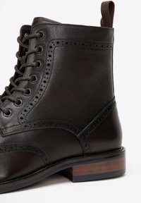 Hnědá kožená kotníková bota s brogue zdobením, kulatými šněrovadly a dřevěným podpatkem. Obsahuje poutko na zadní straně pro snadné obouvání.
