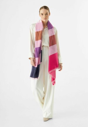 Foulard en blocs de couleurs avec des sections rose, violet, marron et bleu marine ; fabriqué en matériau doux avec des bords à franges ; porté drapé sur une chemise claire.