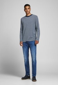Marinblå och vit randig långärmad skjorta, blå jeans och mörka skor. Skjortan har en rund halsringning; jeansen har en något urblek look.