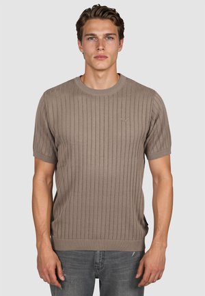 Junger Mann, der einen beige gerippten Kurzarmpullover und graue Jeans trägt, steht vor einem schlichten hellen Hintergrund.