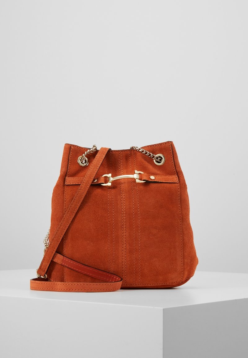 Sac seau en daim couleur orange brûlé avec un haut à cordon, des accents métalliques et une bandoulière amovible. Présente des détails cousus sur le devant.