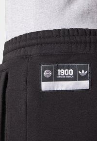 Detailní záběr na pas černých tepláků s logy Bayernu Mnichov a Adidas a našitým nápisem "1900 Bayern Mnichov".