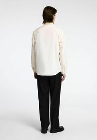 Camicia chiara, con bottoni e vestibilità dritta, indossata con pantaloni e scarpe neri. Il tessuto appare liscio e ben rifinito.