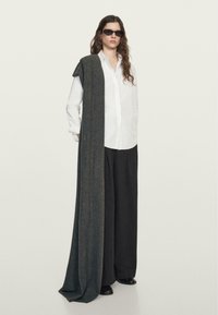 Sciarpa grigia oversize drappeggiata su una camicia bianca con bottoni, abbinata a pantaloni neri larghi e occhiali da sole scuri, creando una silhouette stratificata.