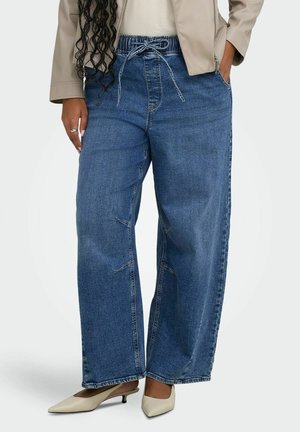 Femme portant un jean en denim bleu à jambes larges avec taille élastique et cordon, des escarpins beige à bout pointu à petit talon, et une veste beige.