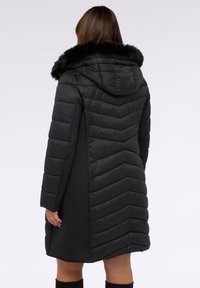 Abrigo de plumas negro con un patrón de flecha, capucha y ribete de pelaje sintético. Presenta un diseño largo y una textura suave para calidez y estilo.