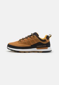 Zapatillas de malla marrones con acentos negros y una suela media blanca. Presenta un upper texturizado, un cuello acolchado y un interior amarillo. Se ve el logotipo de Timberland.