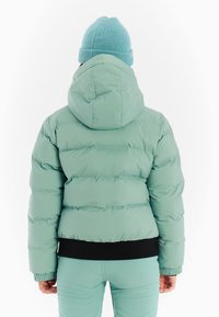 Gevoerde mintgroene jas met een capuchon, zwarte manchetten en een elastische zoom. Gemaakt in combinatie met bijpassende mintgroene broek en een blauwe gebreide beanie.