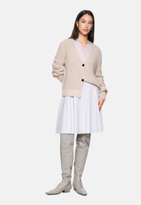 Beige gestrickte Strickjacke mit schwarzen Knöpfen, kombiniert mit einer helllila Bluse und einem weißen Faltenrock. Graue kniehohe Stiefel runden den Look ab.