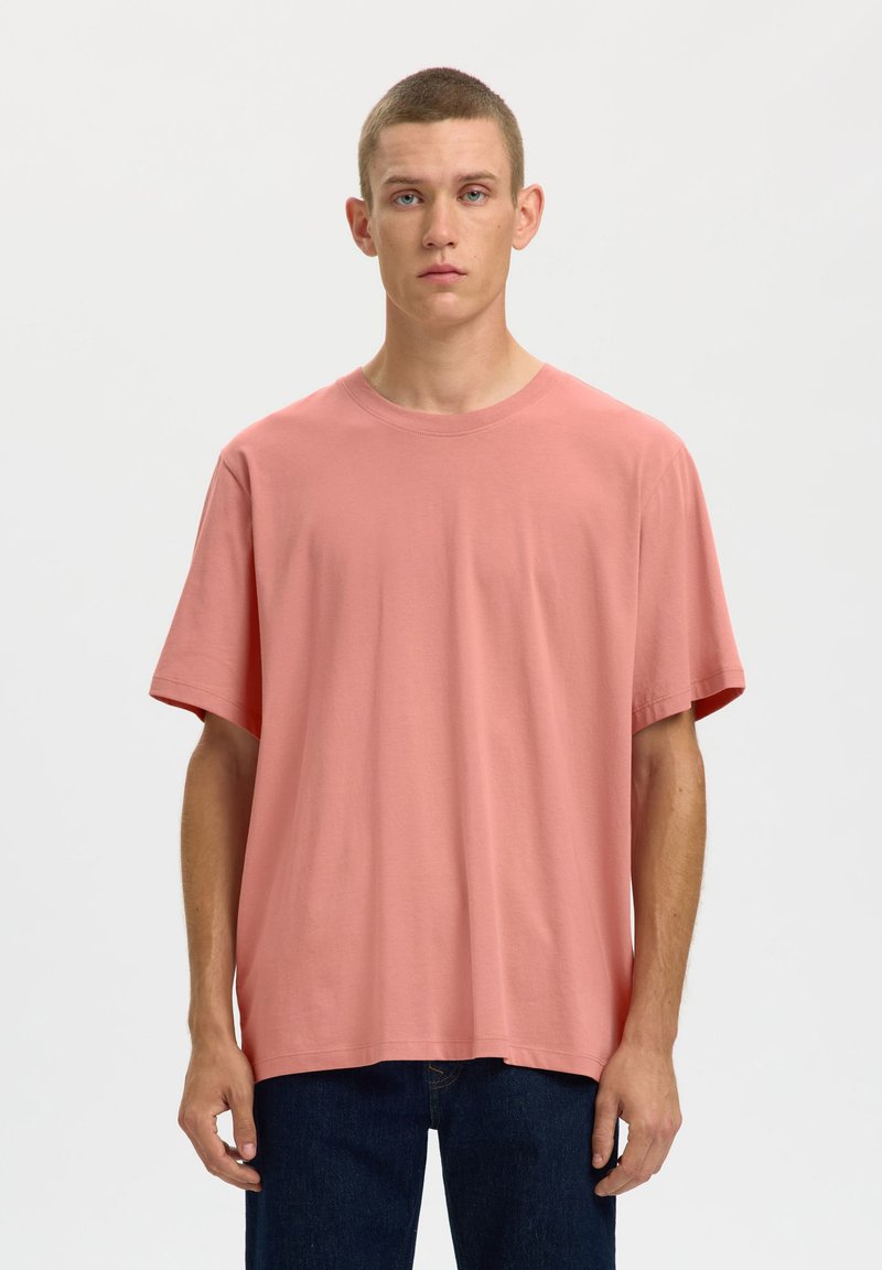 Ανοιχτό ροζ βαμβακερό t-shirt με κοντά μανίκια, στρογγυλή λαιμόκοψη και άνετη γραμμή, φορεμένο με σκούρο μπλε τζιν σε απλό φόντο.