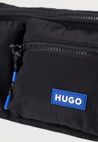 Μαύρη νάιλον τσάντα μέσης με πολλές φερμουάρ τσέπες, με υφή στην επιφάνεια και μπλε ετικέτα με λογότυπο που έχει λευκό κείμενο.