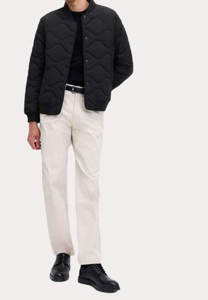 Homme portant une veste bomber matelassée noire, une chemise noire, un pantalon blanc, une ceinture noire et des chaussures en cuir noires, debout devant un fond uni.