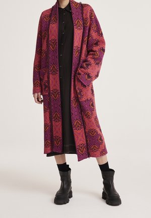 Person, die einen langen gemusterten pink-orangenen Mantel über einem schwarzen Kleid mit schwarzen Stiefeletten trägt und vor einem einfarbigen Hintergrund steht.