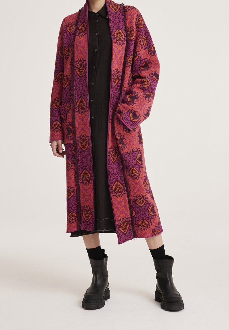 Personne portant un long manteau à motifs roses et orange sur une robe noire avec des bottines noires, debout devant un fond uni.
