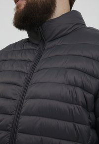 Mørkegrå pufferjakke med quiltet design, høy krage og hel glidelås, med en glatt, glansfull tekstur.