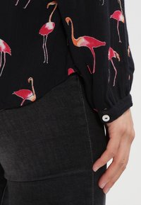 Schwarzes Hemd mit buntem Flamingo-Druck, lange Ärmel und geknöpften Manschetten. Getragen mit dunkelgrauen Hosen. Die Stofftextur wirkt leicht.