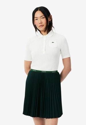 Femme aux longs cheveux foncés porte un polo blanc à manches courtes et une jupe plissée vert foncé avec ceinture à logo Lacoste.