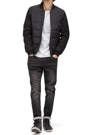 Homme portant une veste bomber matelassée noire sur un t-shirt blanc à rayures, jean noir avec revers roulés, et baskets noires, mains dans les poches.