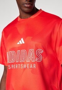 Punainen verkkokangas urheilupaita, jossa on pyöreä kaula-aukko, painettu "adidas"-logo ja valkoiset "SPORTSWEAR"-tekstit. Rakenne on teksturoitu hienovaraisilla kuvioilla.